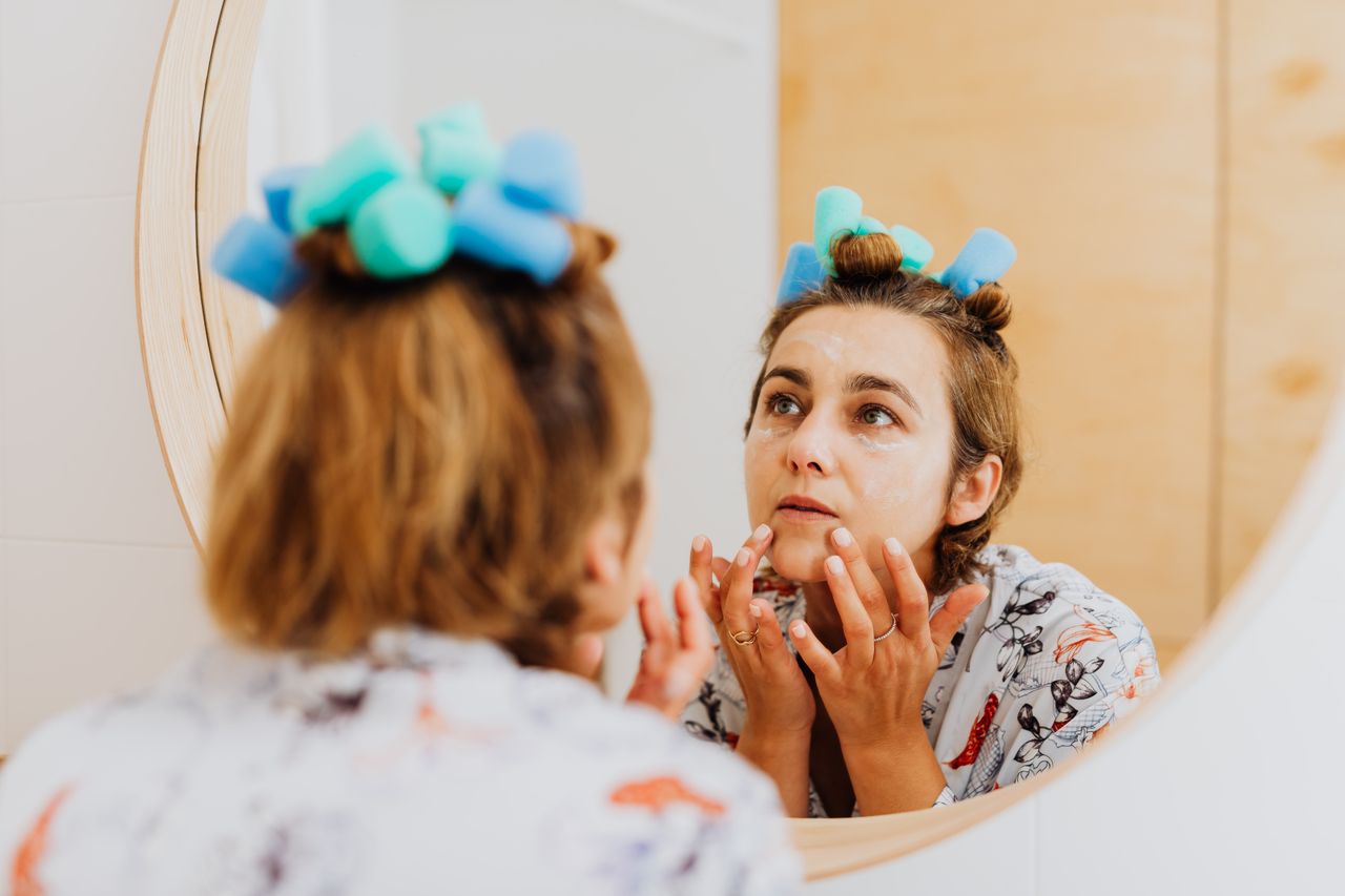 Home Woman with curlers applying cream in front of a mirror indoors.