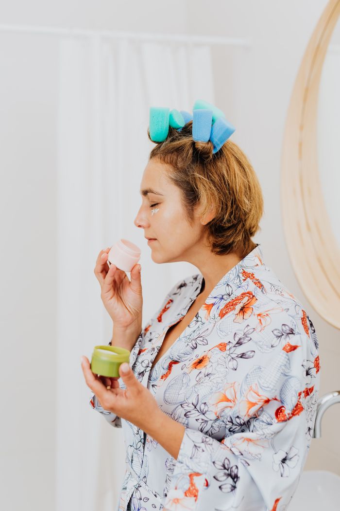 Woman in robe with hair curlers applying face cream in bathroom.