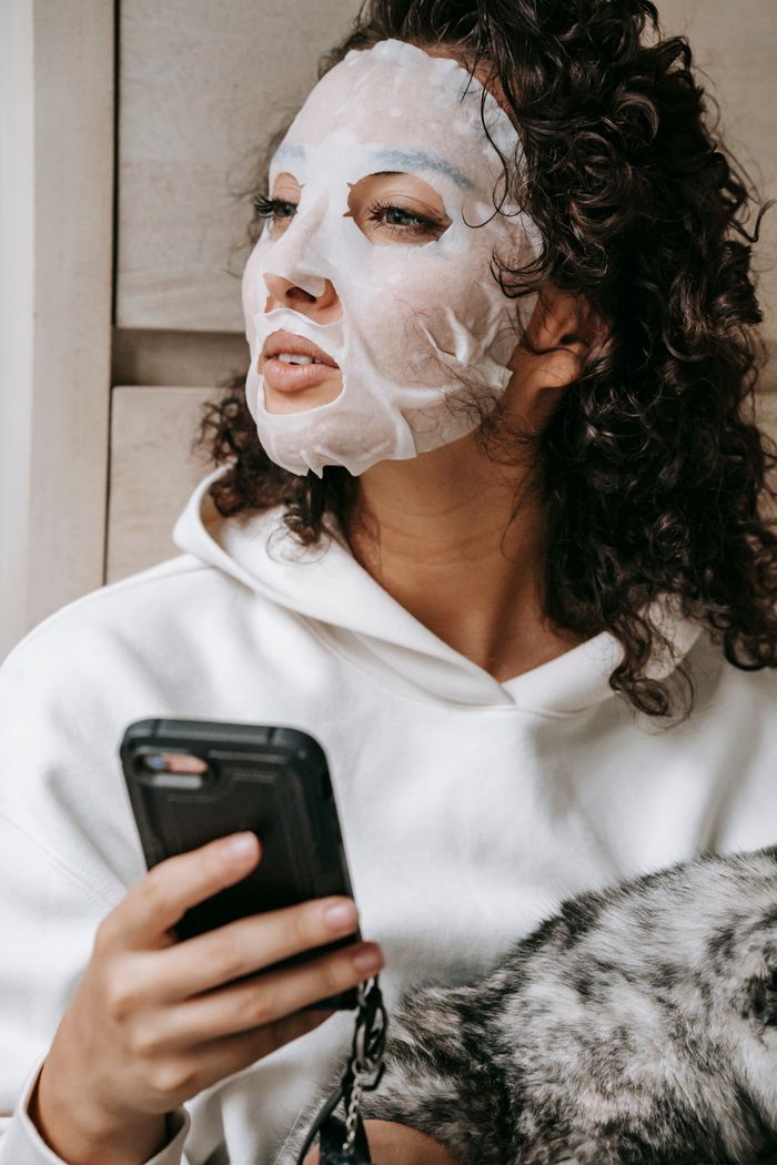 Calm female in sheet mask looking away with interest while sitting with cat and text messaging on cellphone during weekend