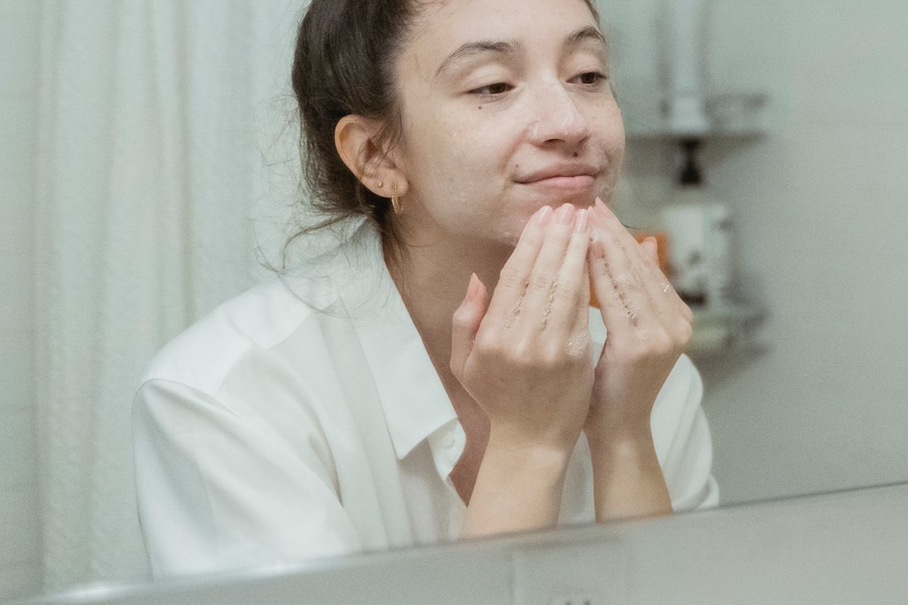 Contact A woman applies facial cream, enjoying her skincare routine in front of a bathroom mirror.