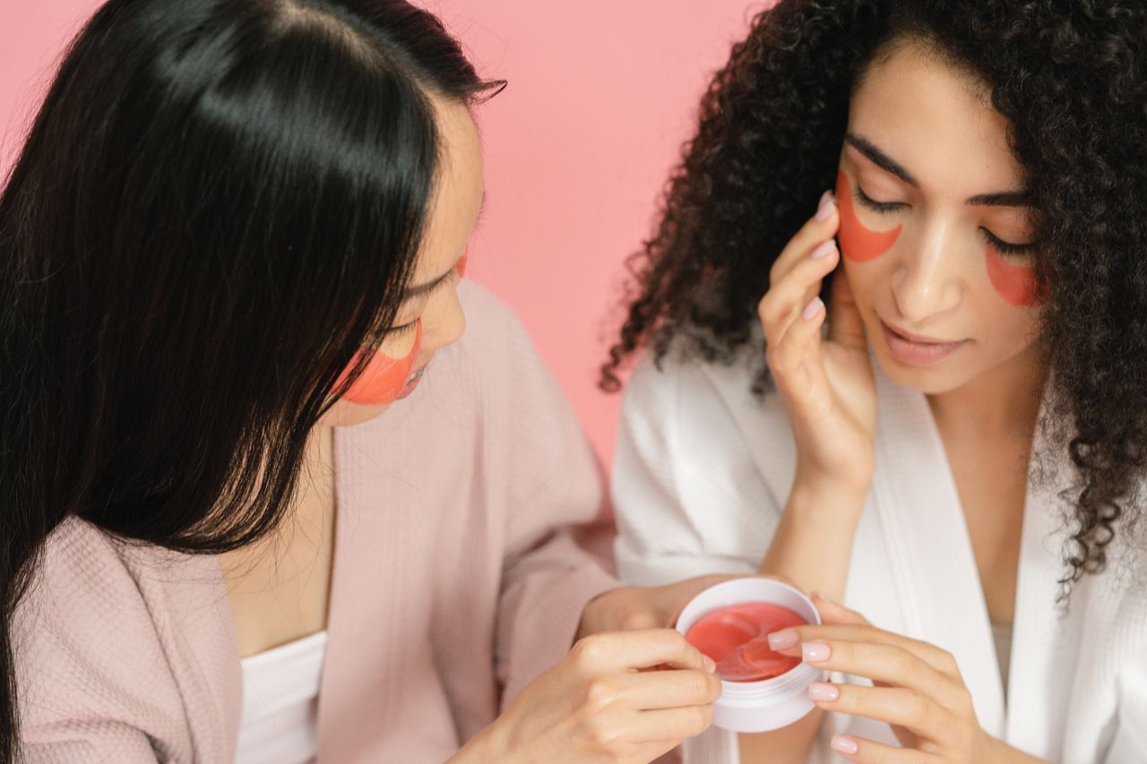 Home Two women applying red eye patches in a skincare routine, focusing on beauty and self-care.